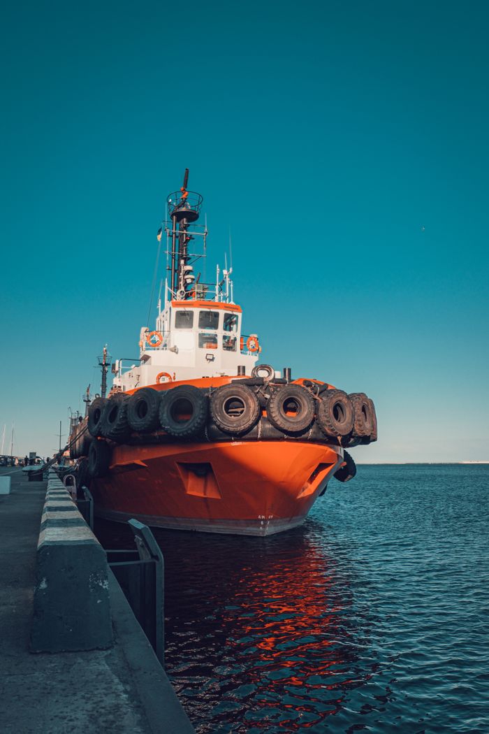 A red tugboat is docked at a pier under a clear blue sky, reflecting in the calm water.